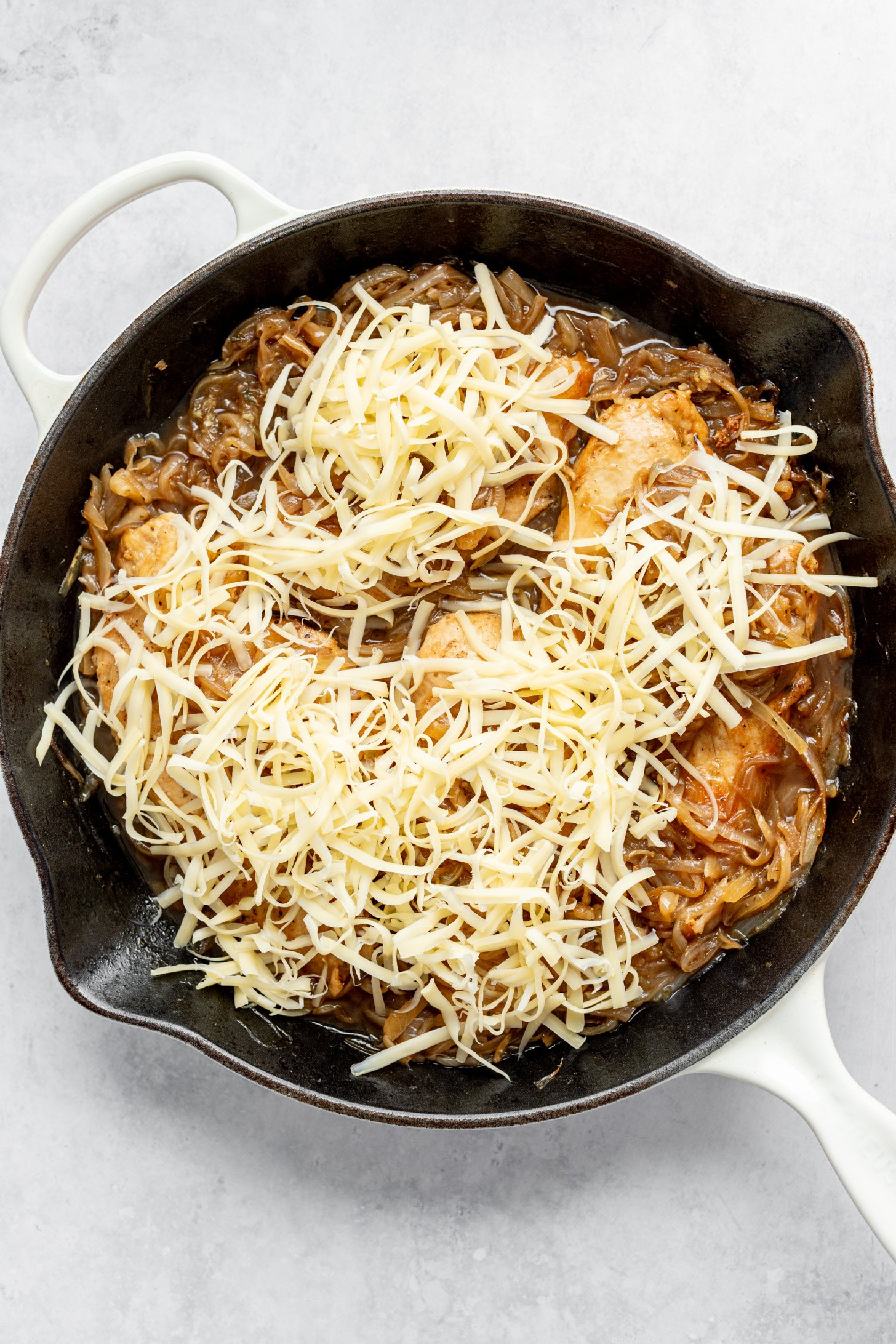 A cast iron skillet filled with caramelized onions, cooked chicken breast and topped with shredded cheese (not yet melted). The skillet is sitting on a countertop.