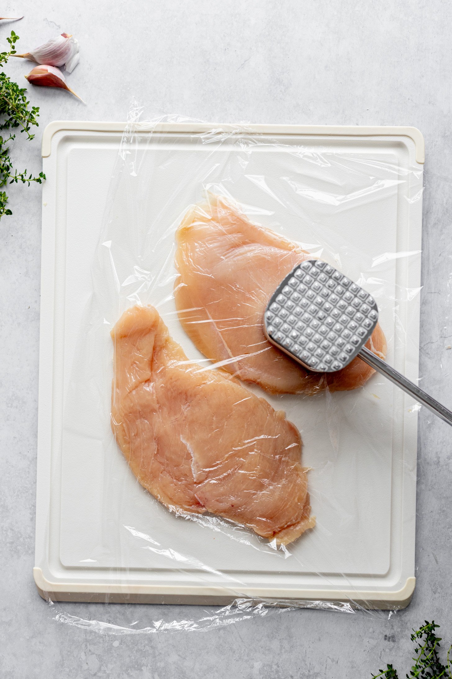 Two chicken breast halves with plastic wrap on top are being pounded flat with a meat tenderizing tool. The chicken breasts are on a cutting board on a counter. The cuttign board is surrounded by a few garlic cloves and fresh herbs.