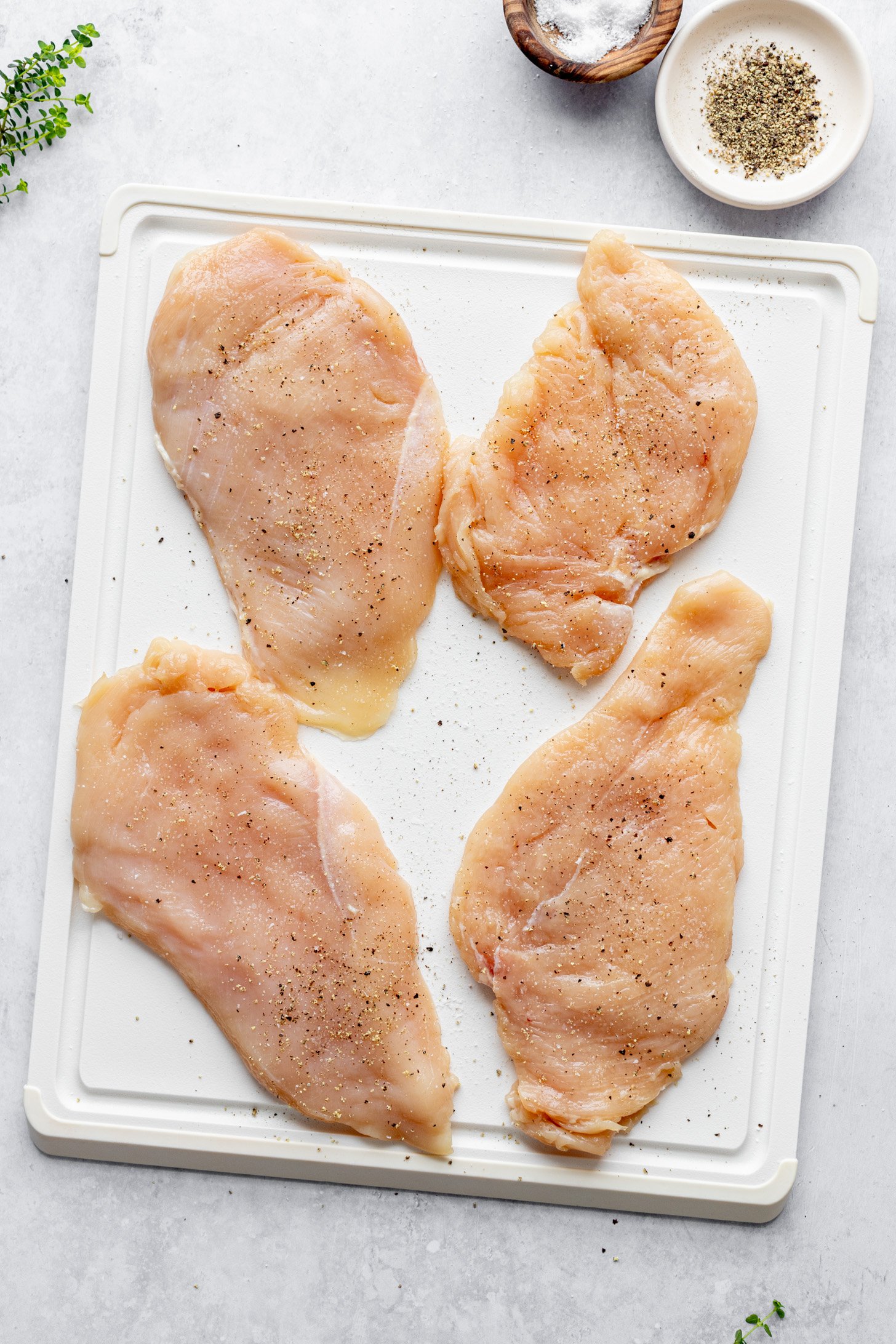 Four raw, flattened chicken breast halves seasoned with salt and pepper on a cutting board. The cutting board is sitting on a counter surrounded by a small bowl of salt, a small bowl of ground black pepper, and some fresh herbs.