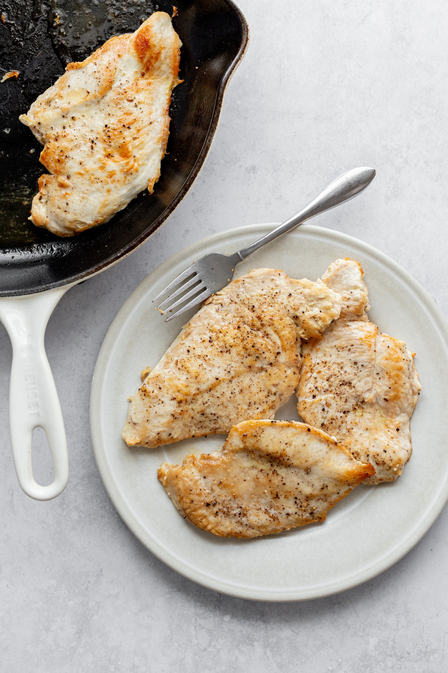 A cast iron skllet with a piece of cooked, seasoned chicken breast is sitting next to a plate filled with 3 pieces of cooked, seasoned chicken breasts. A fork is on the plate and the plate and skillet are sittigng on a countertop.