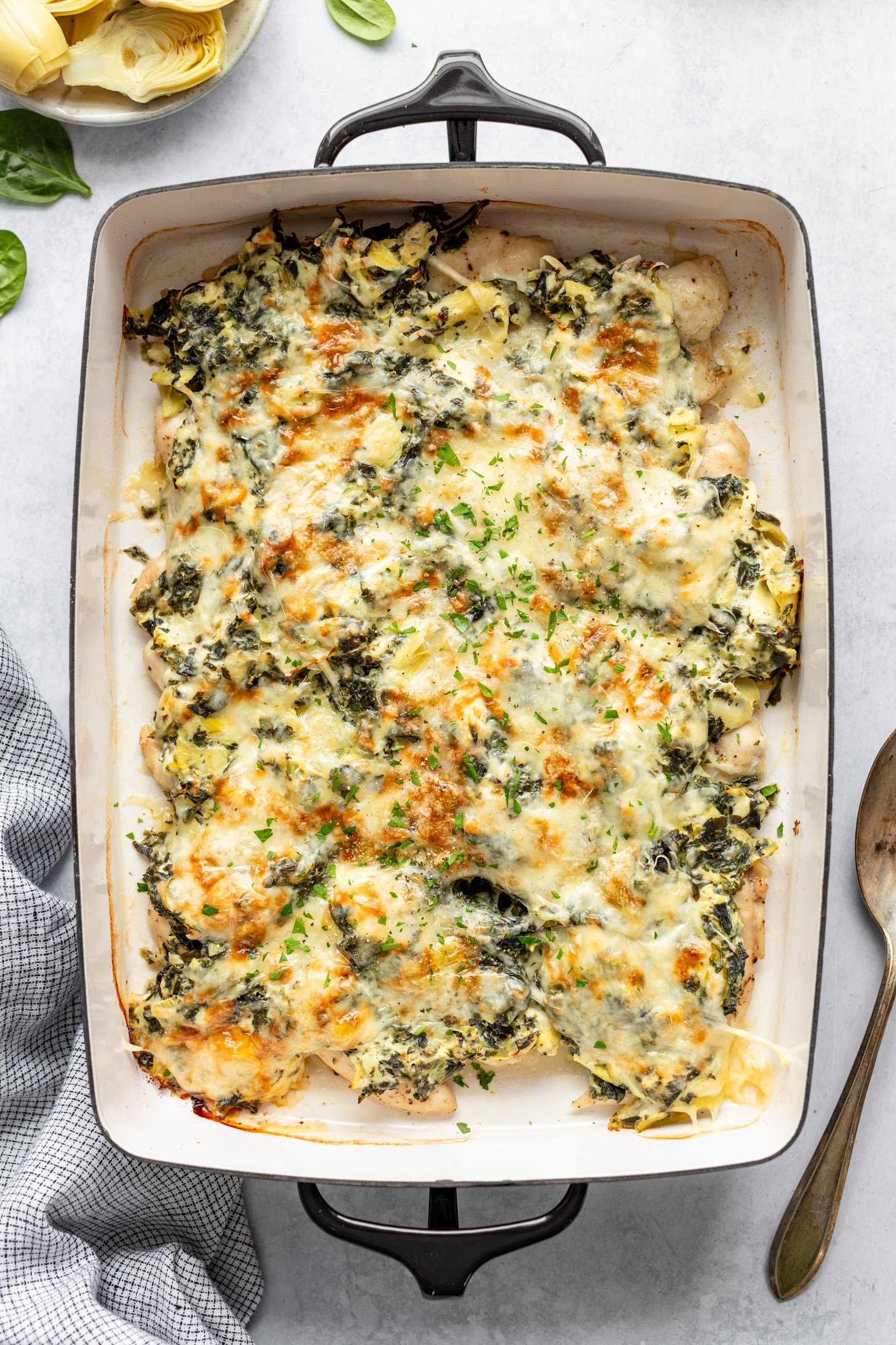 Casserole dish filled with cooked Creamy Chicken Spinach Artichoke Bake sitting on countertop. Casserole dish is surrounded by a napkin, serving spoon, a small dish containing artichoke hearts, and a few spinach leaves strewn about on countertop.