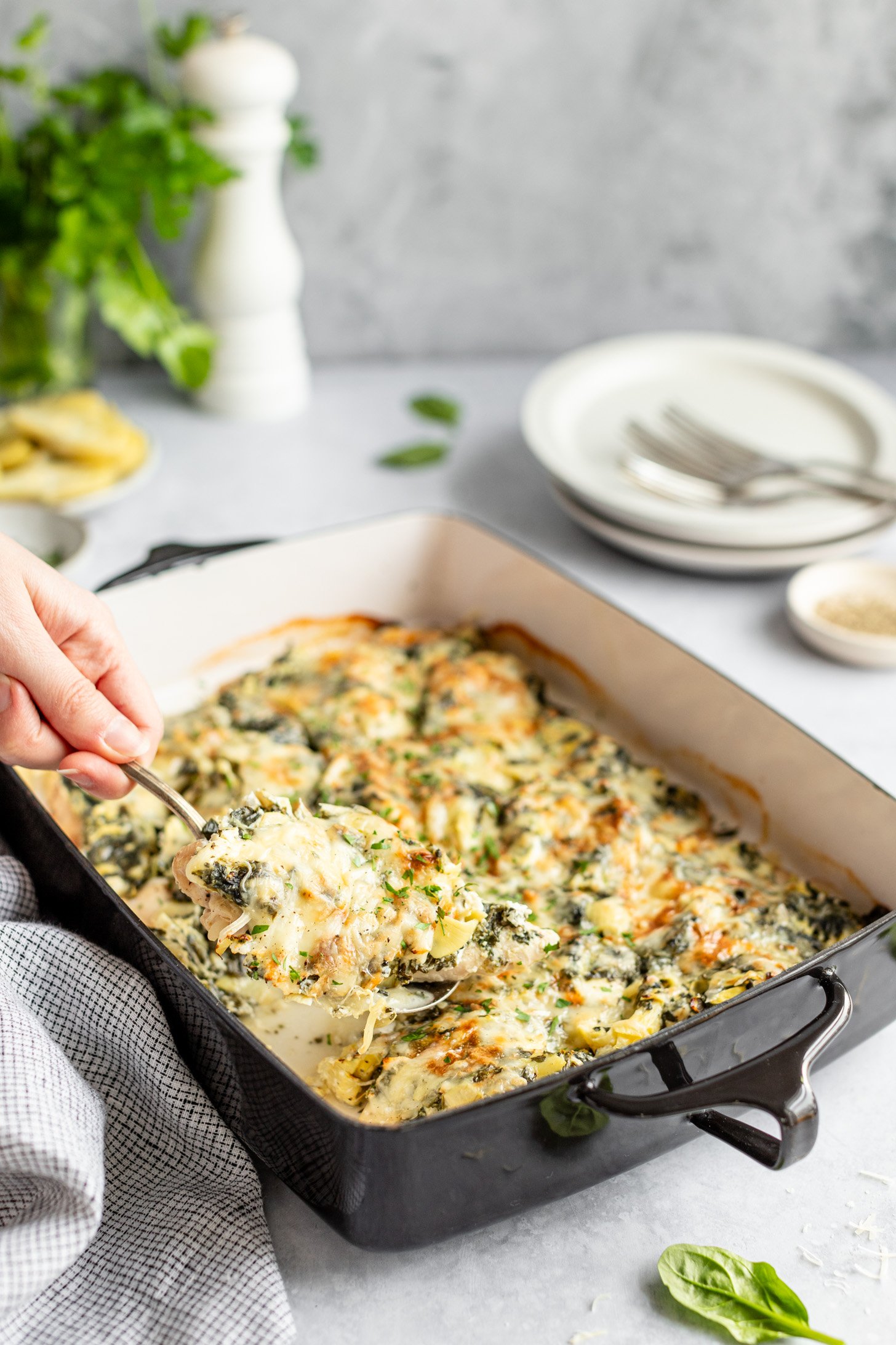 A hand holding a spoon taking a helping of Creamy Chicken Spinach Artichoke Bake out of a casserole dish. Casserole dish is sitting on a countertop and surrounded by a napkin, a few empty dishes topped with forks, small dishes containing various ingredients and some Parmesan cheese and spinach leaves spilled on countertop.