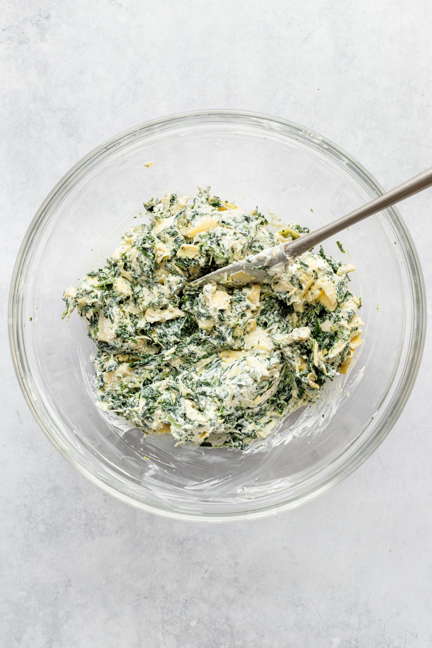 Chopped artichoke hearts, defrosted frozen spinach, Parmesan cheese, garlic, onion powder, salt, black ground pepper, cream cheese, and yogurt all mixed up with a spoon in a glass bowl. Glass bowl is sitting on countertop.