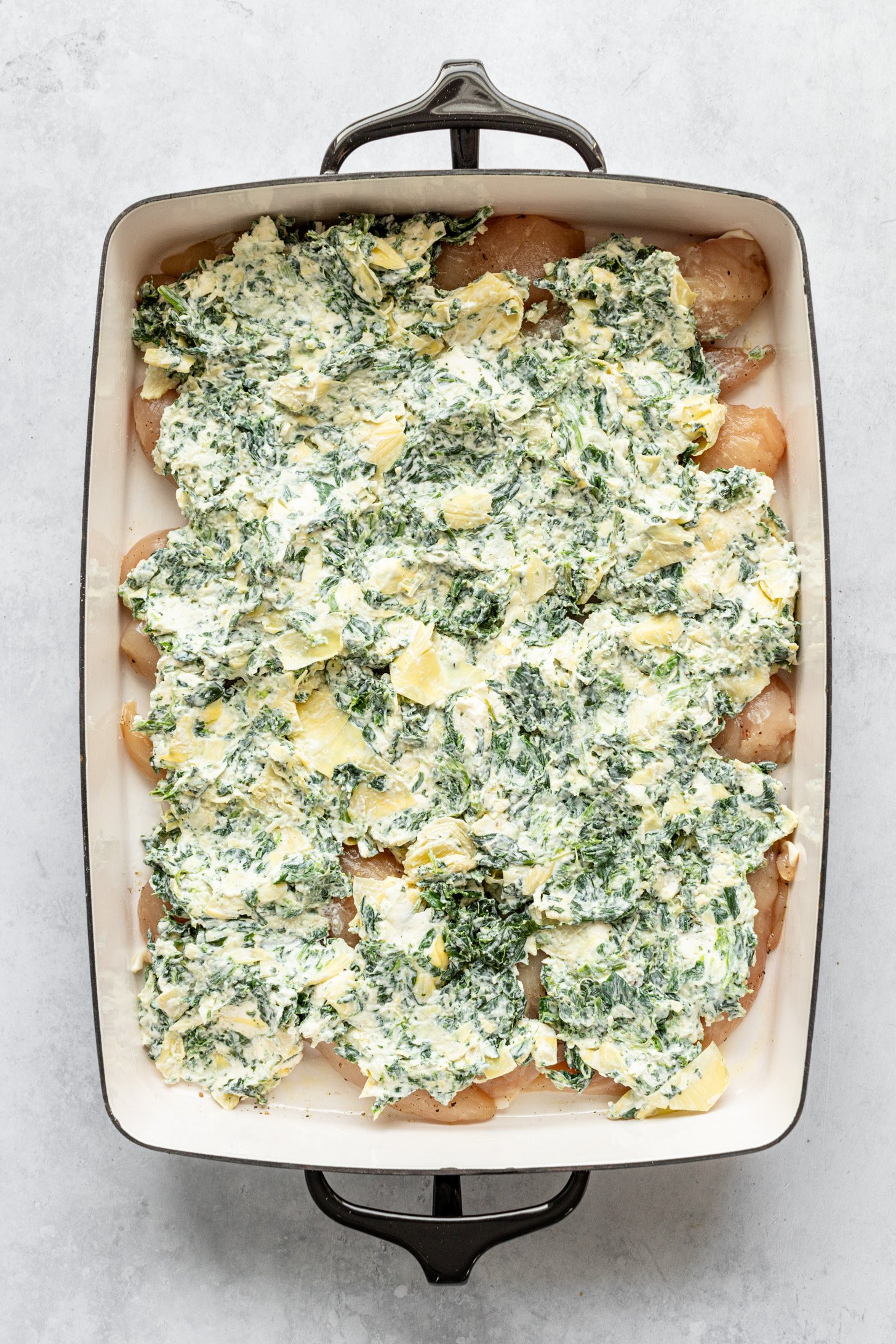 A casserole dish filled with raw chcken breasts topped with cream cheese mixture spread in an even layer. Casserole dish is sitting on a countertop.
