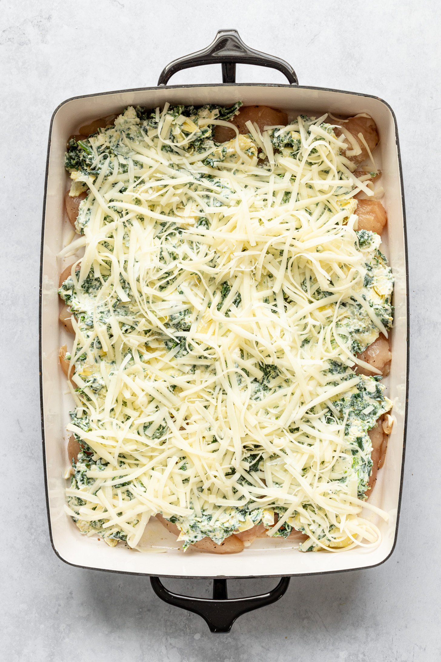 A casserole dish filled with raw chcken breasts topped with a layer of cream cheese mixture and a layer of shredded Parmesan cheese on top. Casserole dish is sitting on a countertop.