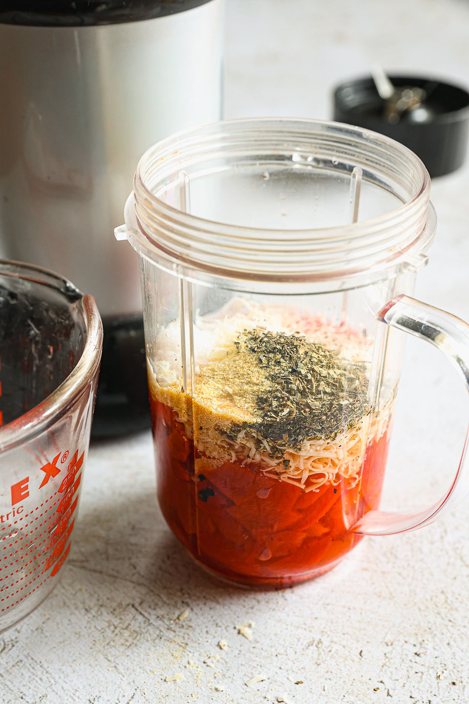 A blender cup filled with ingredients for red pepper sauce prior to blending sits on the counter. Next to the blender cup sits a blender attachment, blender base, and a glass measuring cup.