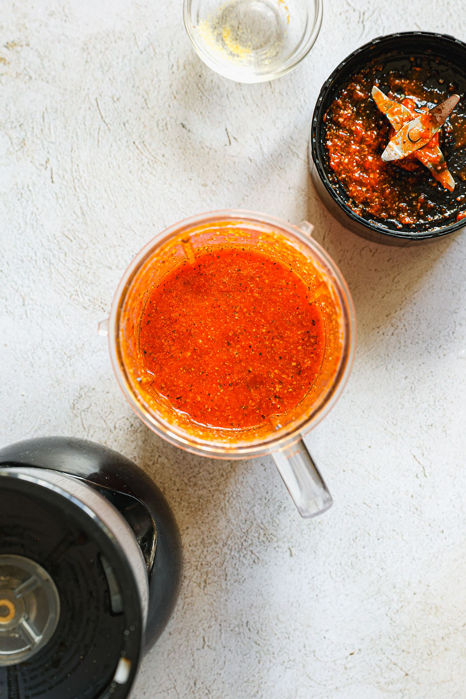A blender cup filled with blended ingredients for red pepper sauce sits on a counter. Next to the blender cup sits the blender base, a blender blade attachment, and a small glass dish.