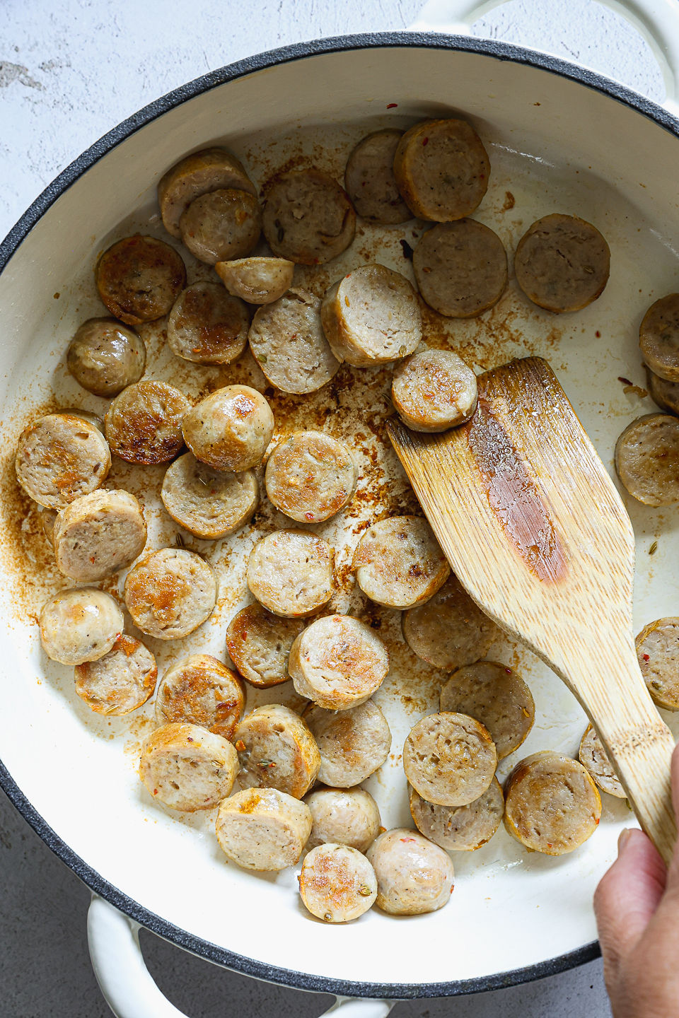 Slices of sausage being sautéed in a pot using a wooden spoon. The pot sits on a countertop.