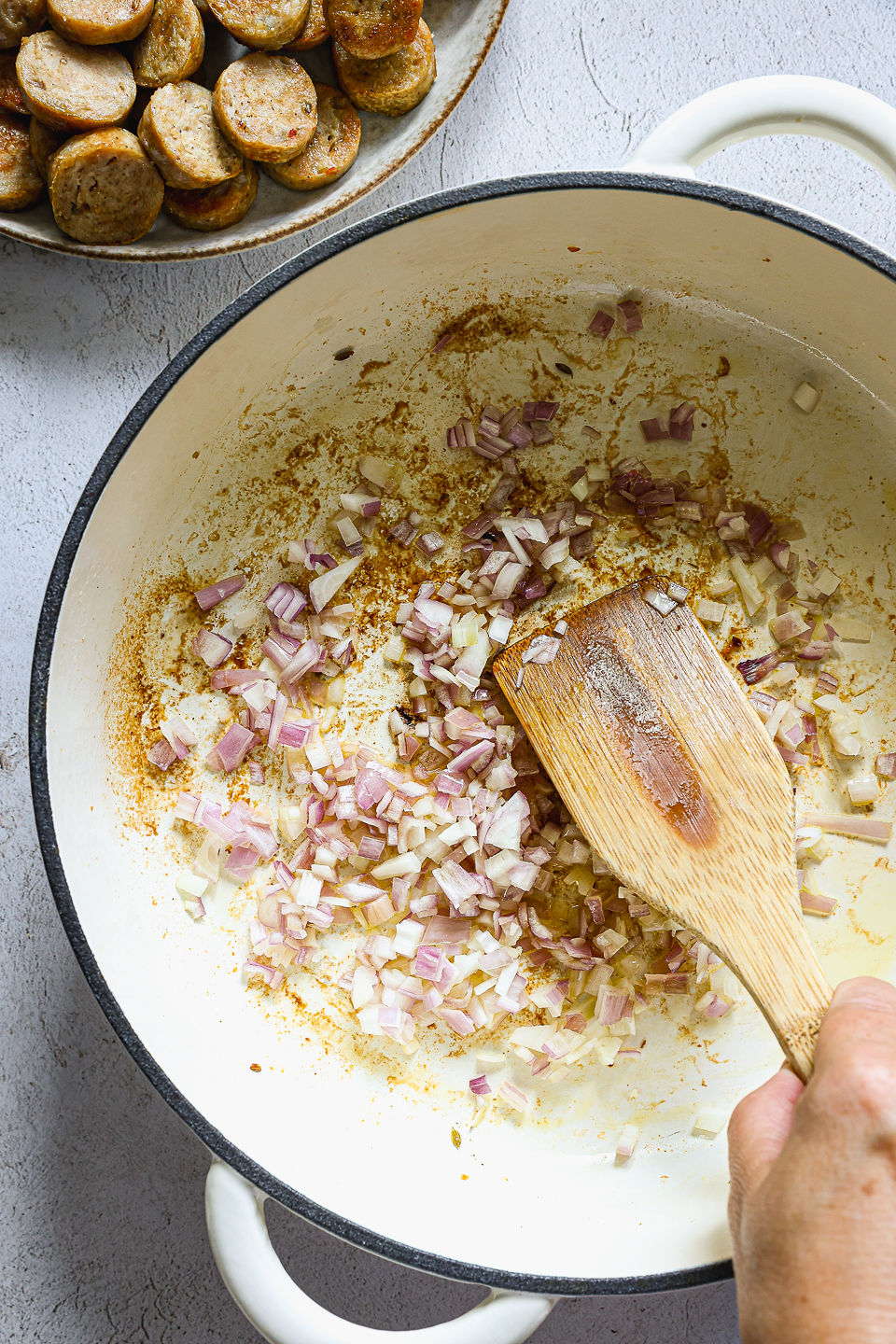 Shallots being sautéed in a pot using a wooden spoon. Next to the pot on the countertop sits a bowl of sliced sausage.