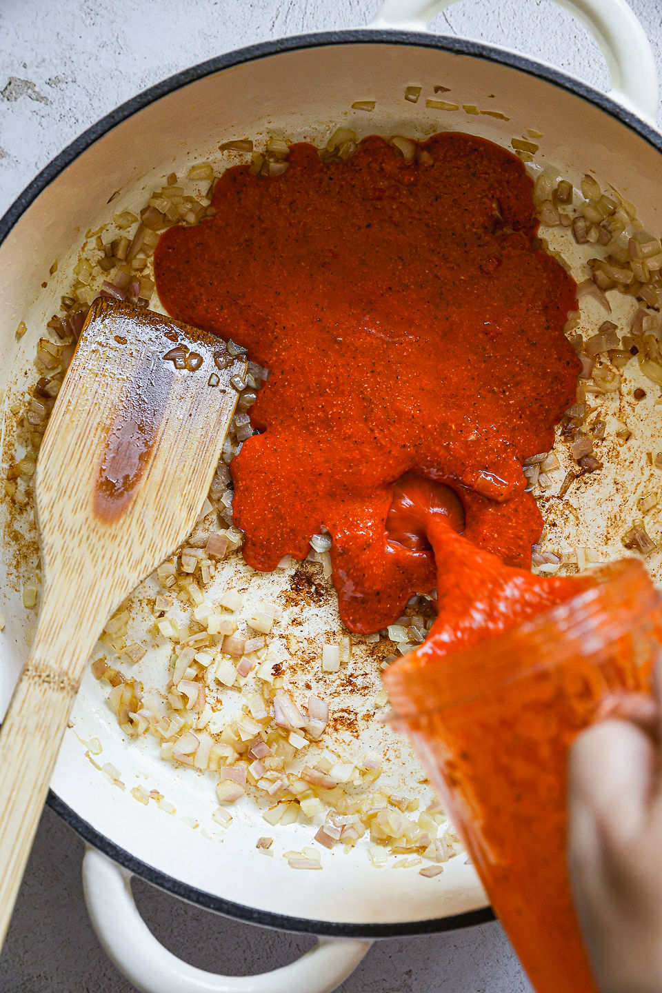 Blended red pepper sauce being poured by hand from a blender cup into a pot with sautéed shallots. A wooden spoon sits in the pot. The pot sits on a countertop.