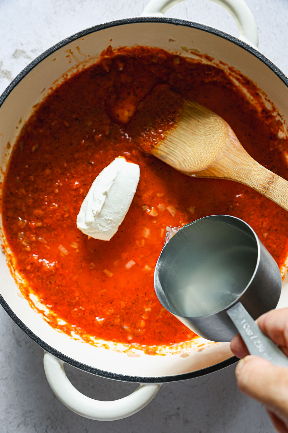 A hunk of cream cheese sits in a pot of red pepper sauce prior to being mixed in. Broth is being poured from a metal measuring cup into the pot. A wooden spoon sits in the pot, and the pot sits on the countertop.