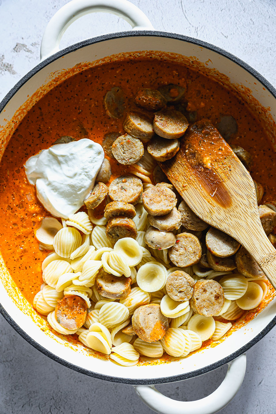 A hunk of cream cheese, cooked orecchiette pasta, and sliced sausage sit in a pot of red pepper sauce prior to being mixed in. A wooden spoon sits in the pot, and the pot sits on the countertop.