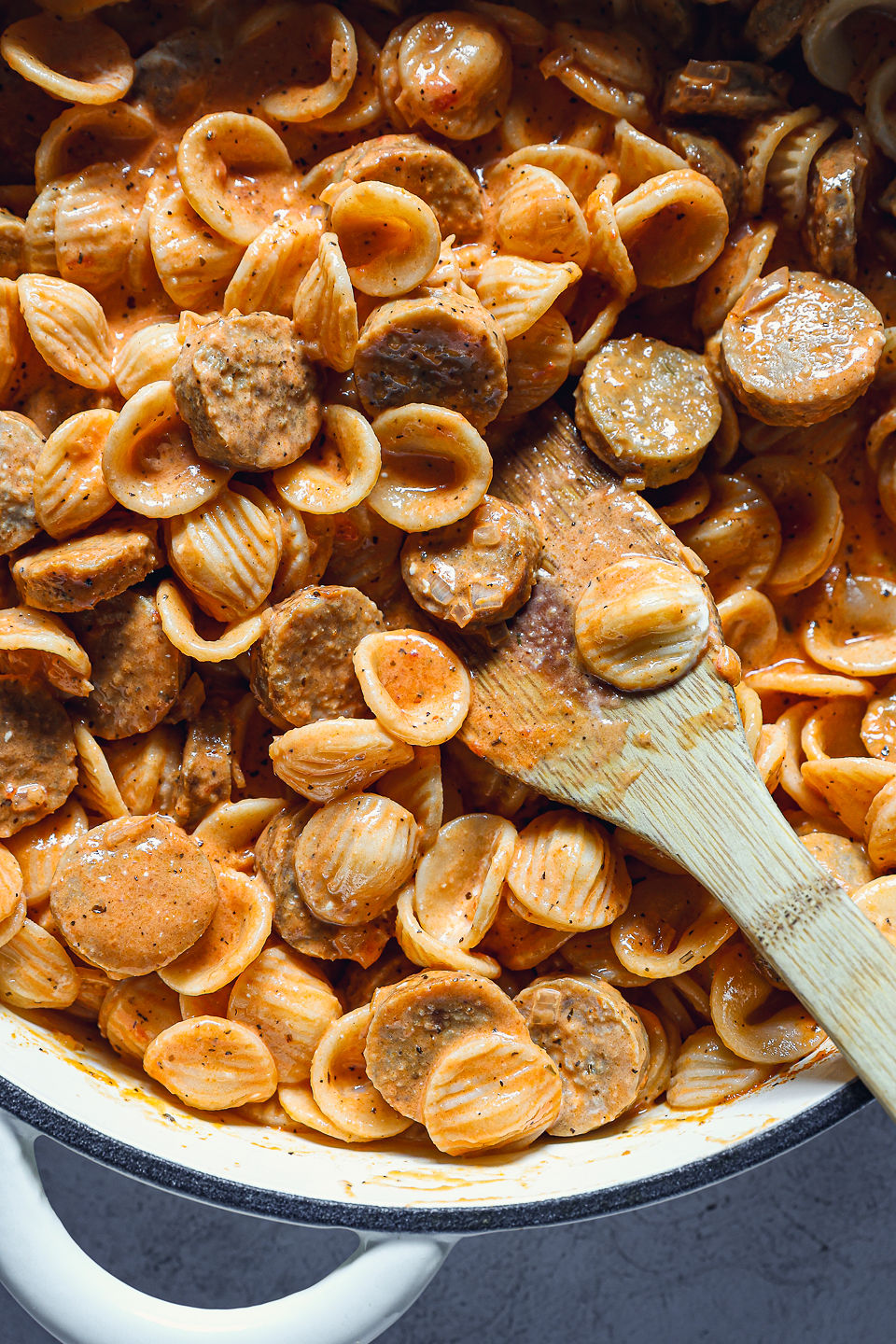 Cooked orecchiette and sausage slices covered in creamy red pepper sauce. The pasta is in a pot, and a wooden spoon sits in the pasta.