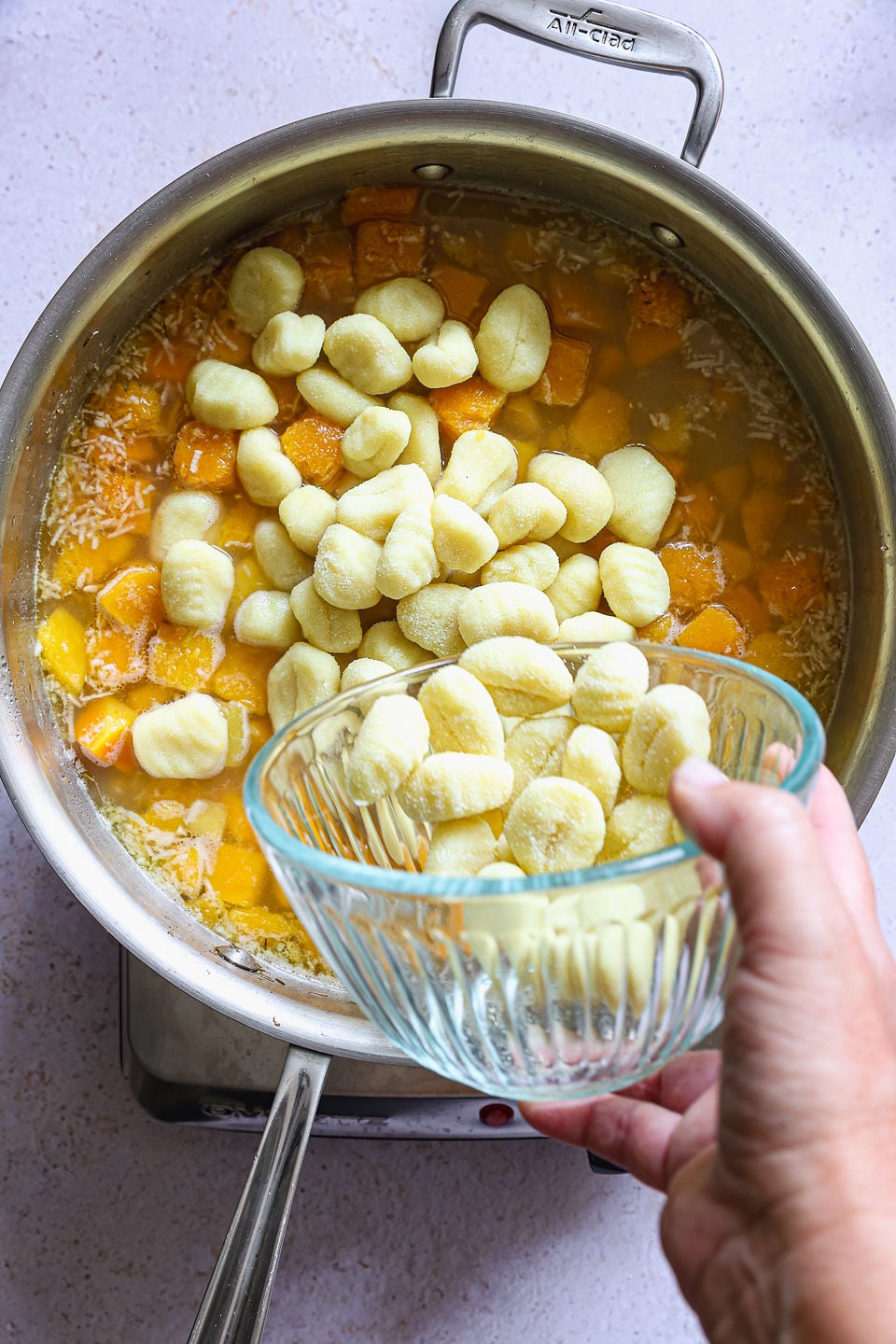 A hand holds a glass bowl of raw butternut squash gnocchi over a stainless steel pot filled with broth and diced butternut squash, preparing to add the dumplings to the simmering liquid.