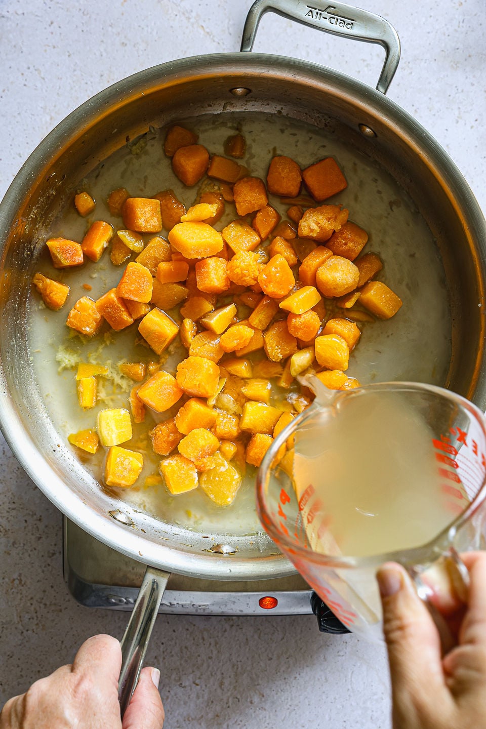 A hand pours liquid from a measuring cup into a skillet with saut&eacute;ed orange cubed vegetables, possibly butternut squash or sweet potatoes, on a stovetop&mdash;perfectly preparing the flavorful base for homemade butternut squash gnocchi.
