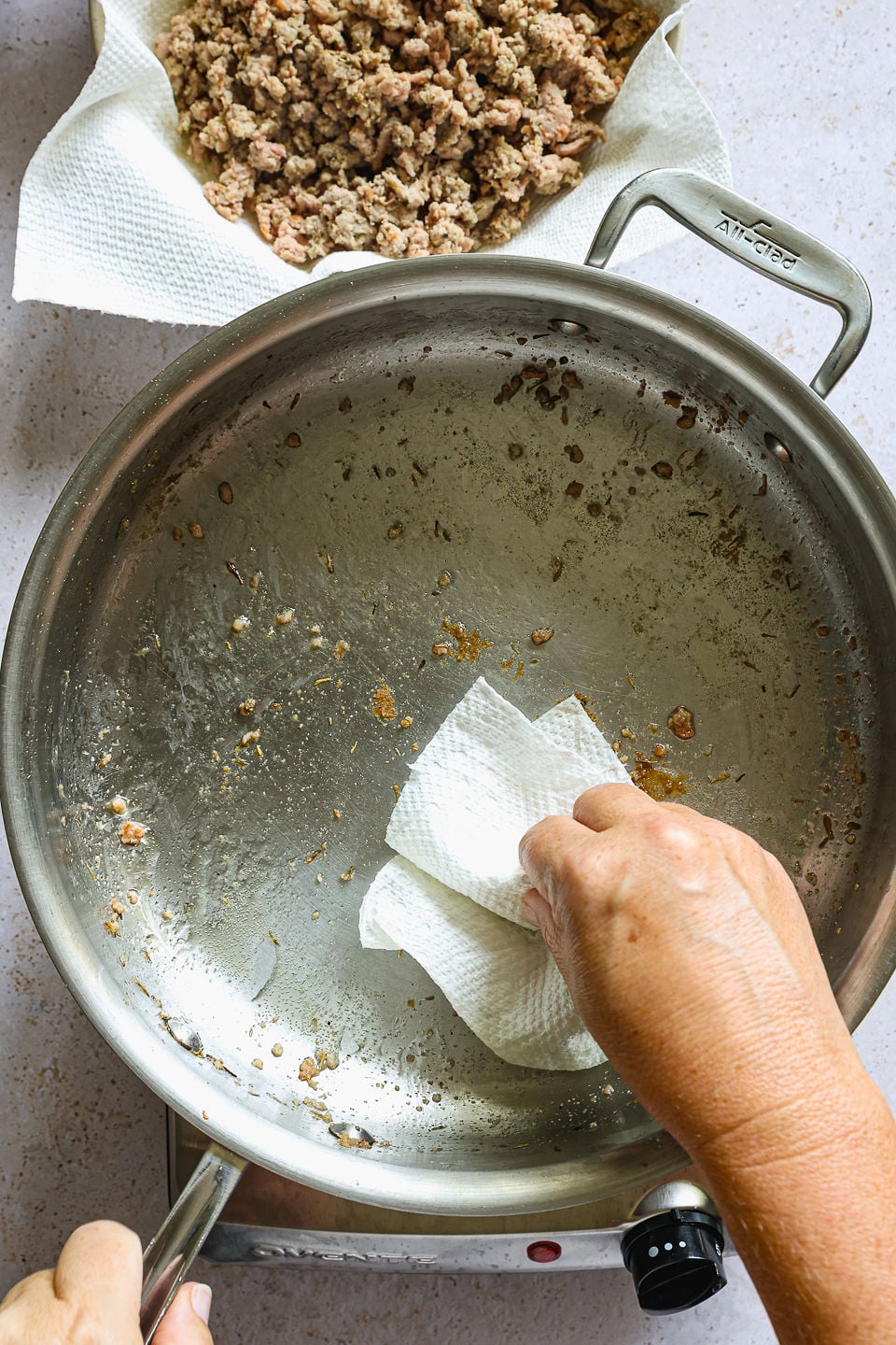 A top-down view shows a hand using a paper towel to wipe grease from a stainless steel pan, prepping the workspace for the next step in making butternut squash gnocchi. Nearby, a separate bowl contains cooked, browned ground meat resting on a paper towel.