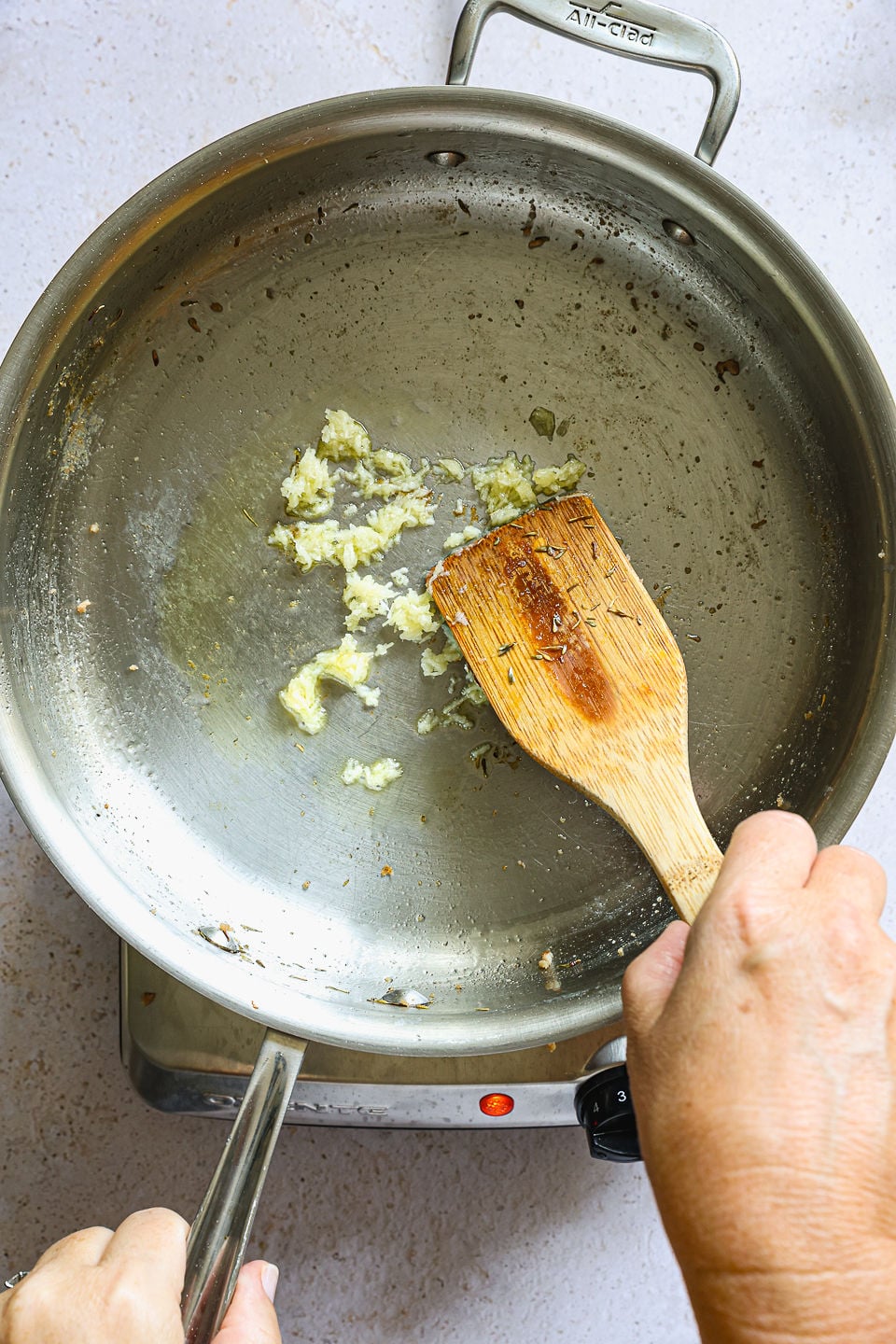A person saut&eacute;ing minced garlic in a large stainless steel pan on a stovetop, using a wooden spatula to stir the garlic in preparation for butternut squash gnocchi.
