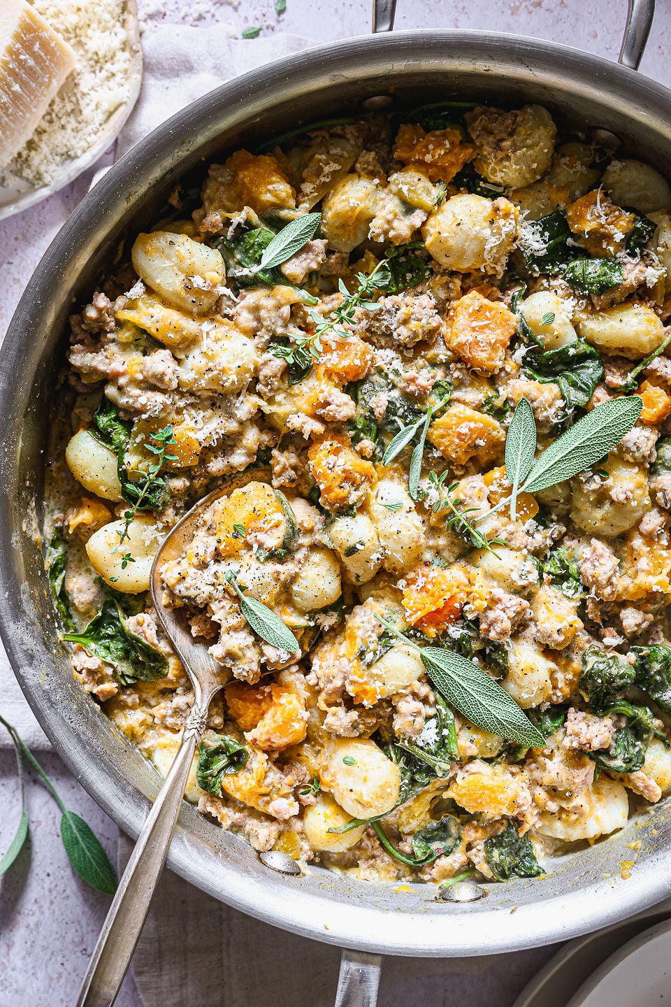 A stainless steel skillet filled with butternut squash gnocchi, ground sausage, roasted butternut squash, and wilted spinach in a creamy sauce. A spoon rests in the dish, which is garnished with fresh sage leaves, thyme sprigs, and grated parmesan cheese.