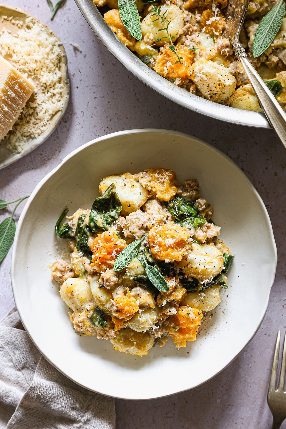 A bowl of butternut squash gnocchi with sausage, spinach, and roasted butternut squash, garnished with sage and grated cheese. A skillet of the same butternut squash gnocchi dish and a plate of cheese are nearby.
