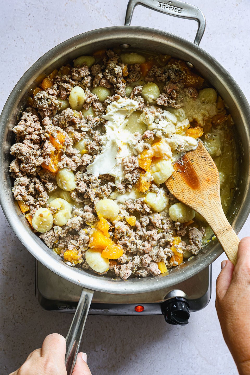 A person stirs a pot of ground meat, butternut squash gnocchi, orange squash pieces, and cream cheese with a wooden spoon on a stovetop.
