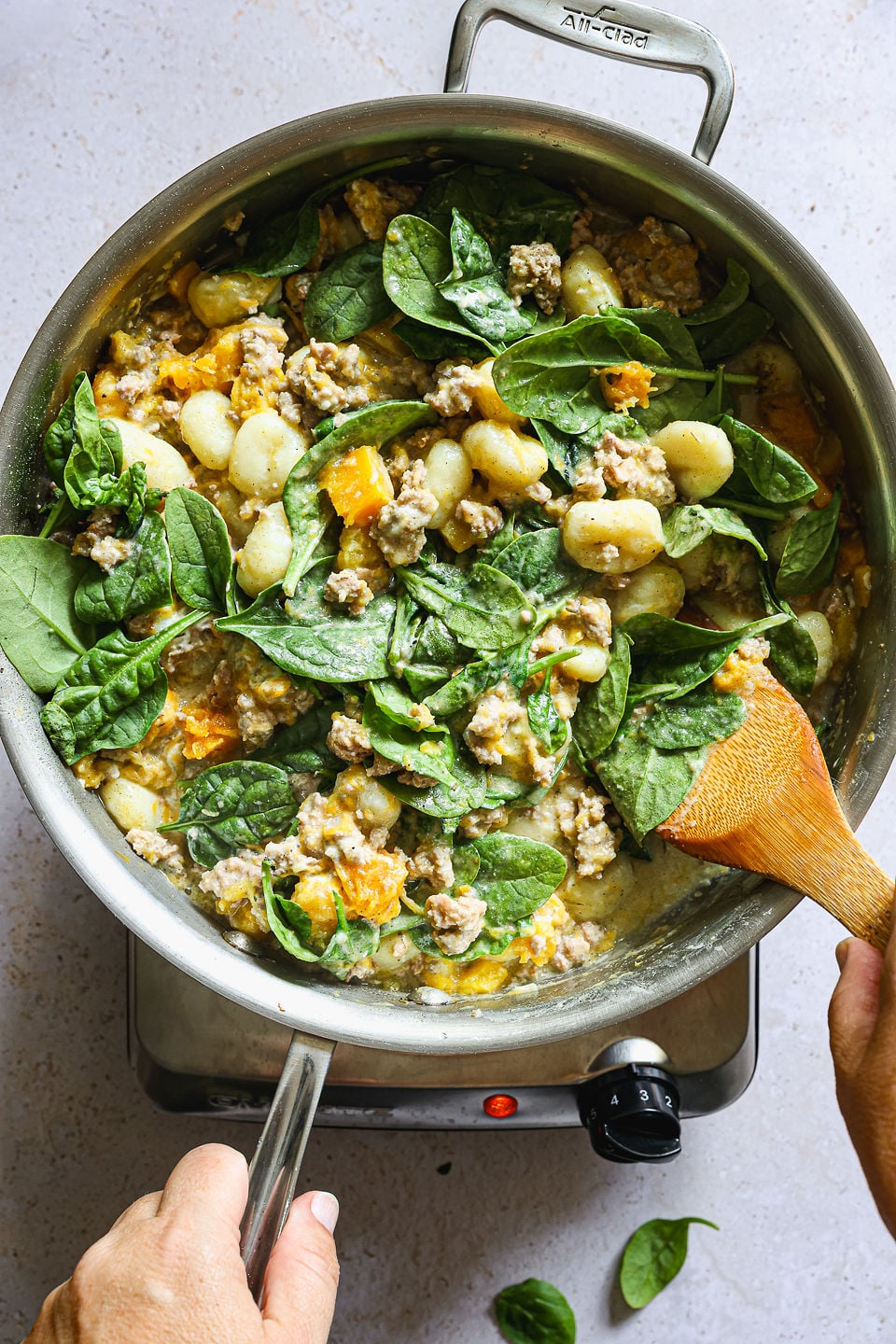 An overhead shot of a large metal skillet on a burner, filled with a mixture of butternut squash gnocchi, ground sausage, roasted butternut squash, and fresh spinach in a creamy sauce, with a wooden spatula mixing the ingredients.