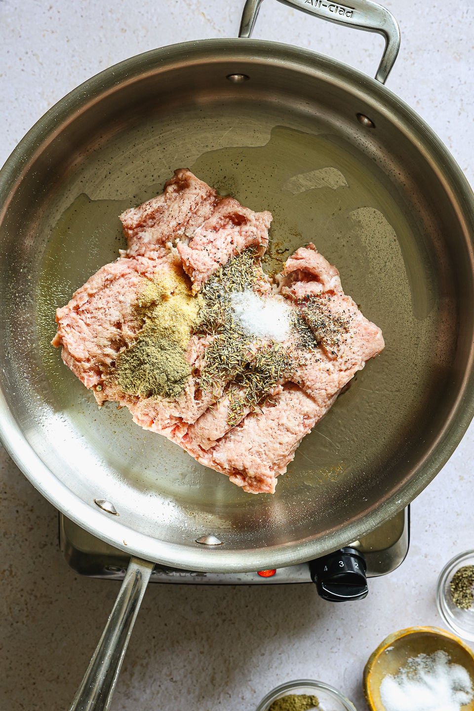 Top-down view of raw ground meat in a stainless steel skillet. The meat is topped with mounds of various seasonings including salt, dried herbs, and spices, resting on a thin layer of oil&mdash;perfect for creating a hearty base to pair with butternut squash gnocchi, ready to be cooked.