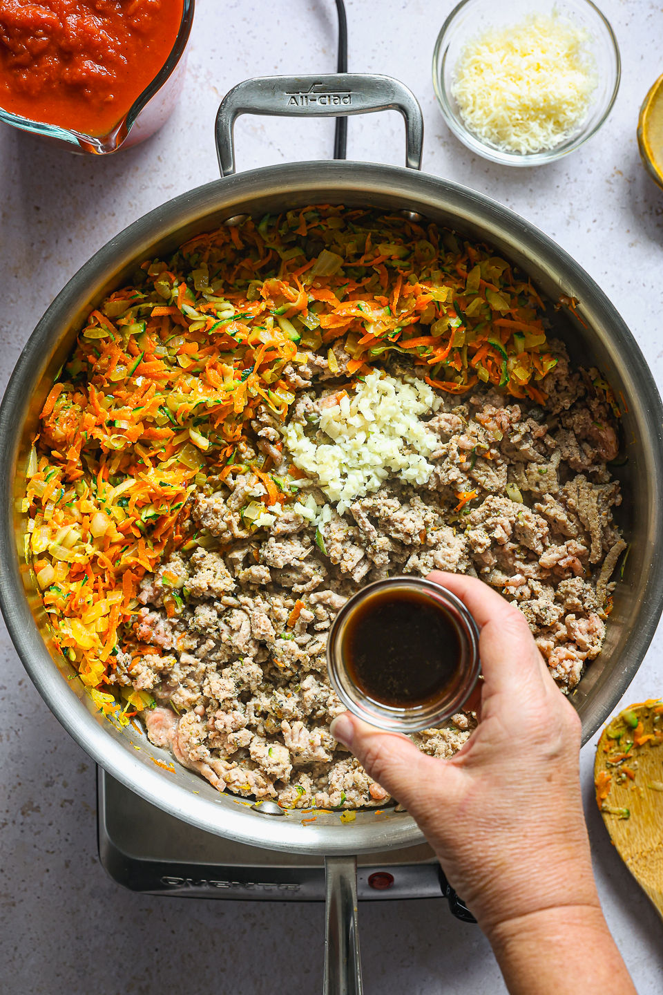 A small container of Worcestershire sauce is being held above a skillet filled with cooked ground turkey, crushed garlic, and a cooked vegetable mixture. The ground turkey, garlic, and vegetable mixture are not yet mixed together. The skillet sits on a single burner on a countertop. Next to the burner sits a used wooden spoon, a small dish of grated Parmesan cheese, and a glass measuring cup of tomato sauce.