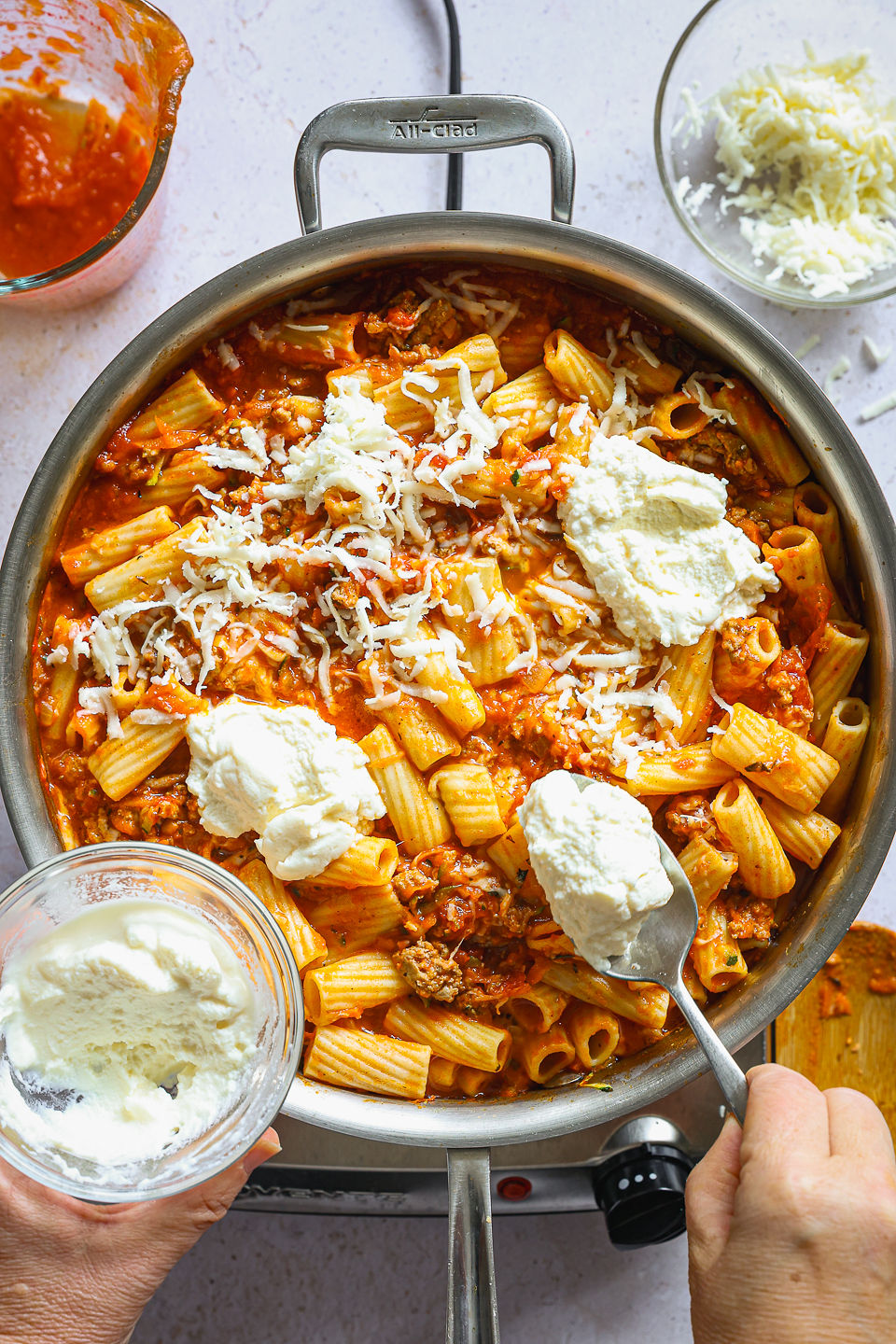 Ricotta is being dropped in dollops using a spoon onto a skilled filled with the cooked, mixed, remaining ingredients for turkey skillet lasagna. A bowl with the remaining ricotta is being held by hand near the skillet. The skillet sits on a single burner on the countertop. Next to the skillet sits a used wooden spoon, a glass measuring cup with some remaining tomato sauce, and a glass dish with shredded mozzarella cheese.