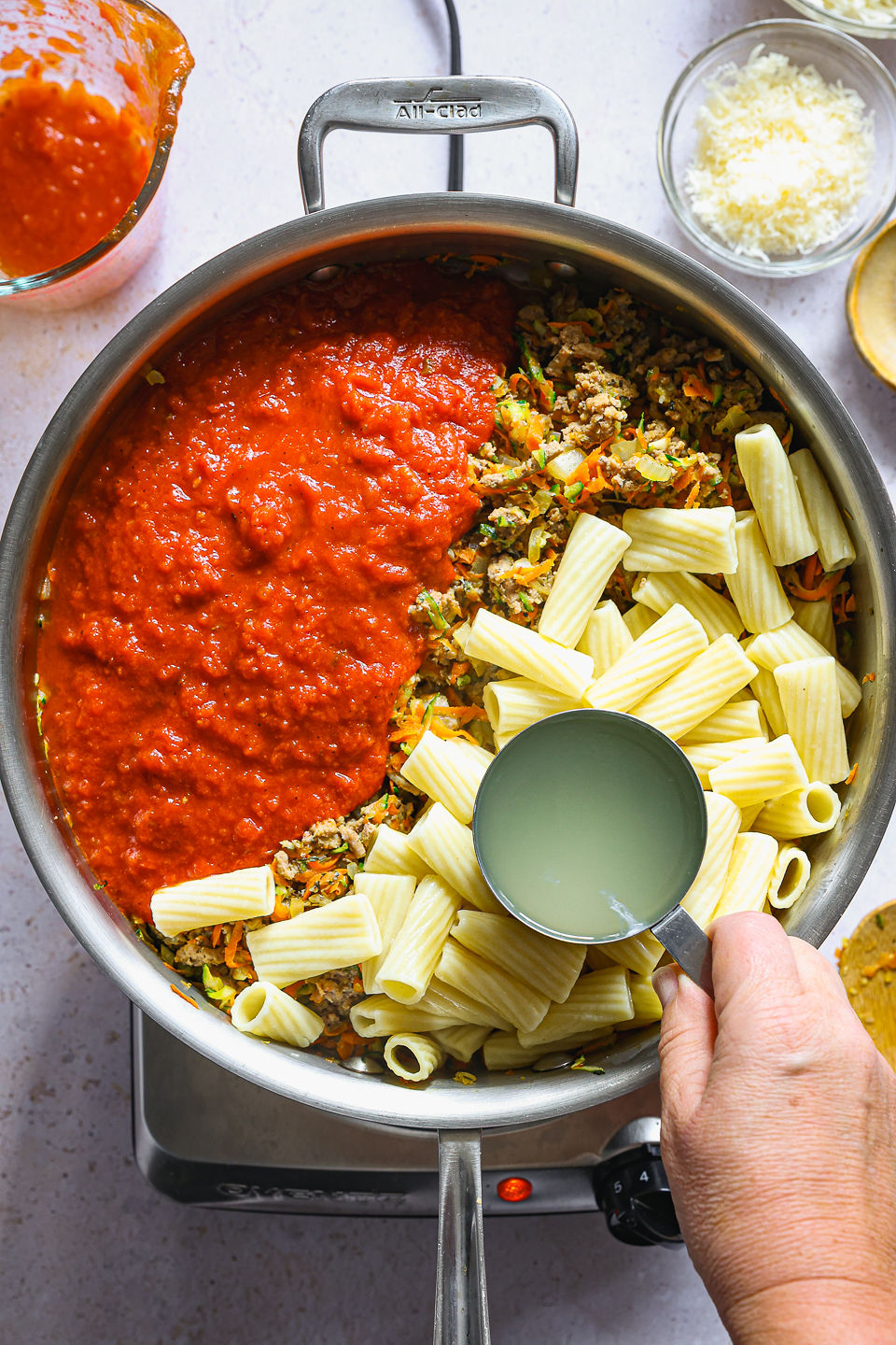 A measuring cup filled with pasta water being held by hand above a skillet of cooked pasta, tomato sauce, cooked ground turkey, and a cooked vegetable mixture, all unmixed. The skillet sits on a single burner on the countertop. Next to the skillet on the countertop sits a used wooden spoon, a measuring cup with some remaining tomato sauce, and a small dish of grated Parmesan cheese.