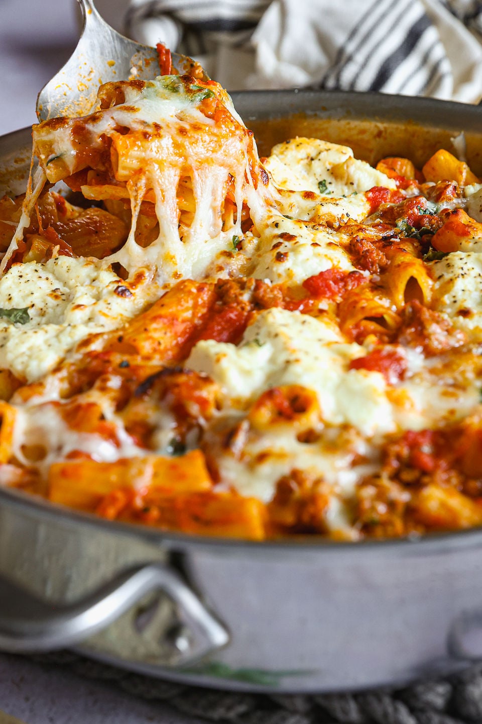 A spoon taking a helping of turkey skillet lasagna from a skillet. The skillet sits on a counter. Next to the skillet sits a striped kitchen towel.
