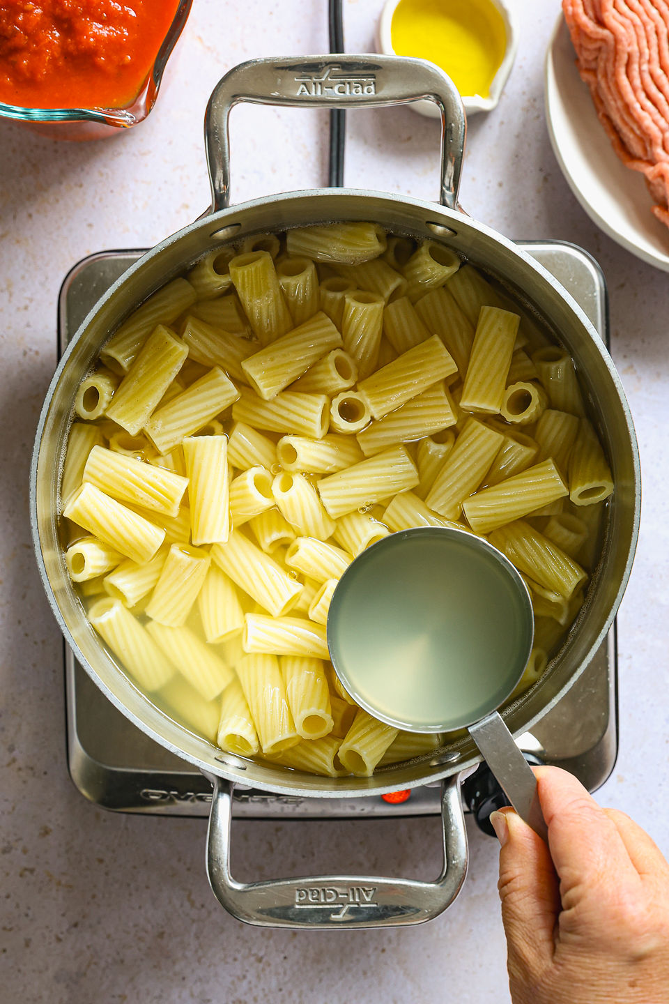 A measuring cup filled with pasta water being held by hand above a large pot filled with cooked pasta in water. The pot sits on a single burner sitting on a countertop. On the counter next to the burner is a uncooked ground turkey on a plate, a small container of olive oil, and a glass measuring cup of pasta sauce.