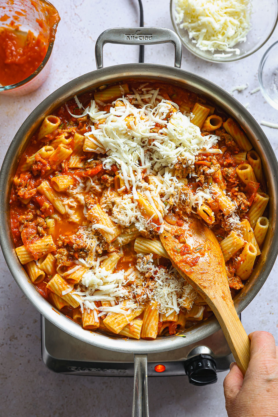 Shredded mozzarella and Grated Parmesan cheese being mixed by hand using a wooden spoon into a mixture of cooked pasta, ground turkey, and vegetable mixture in a skillet. Next to the skillet on the counter sits a glass measuring cup with some remaining tomato sauce and a glass dish filled with shredded mozzarella.