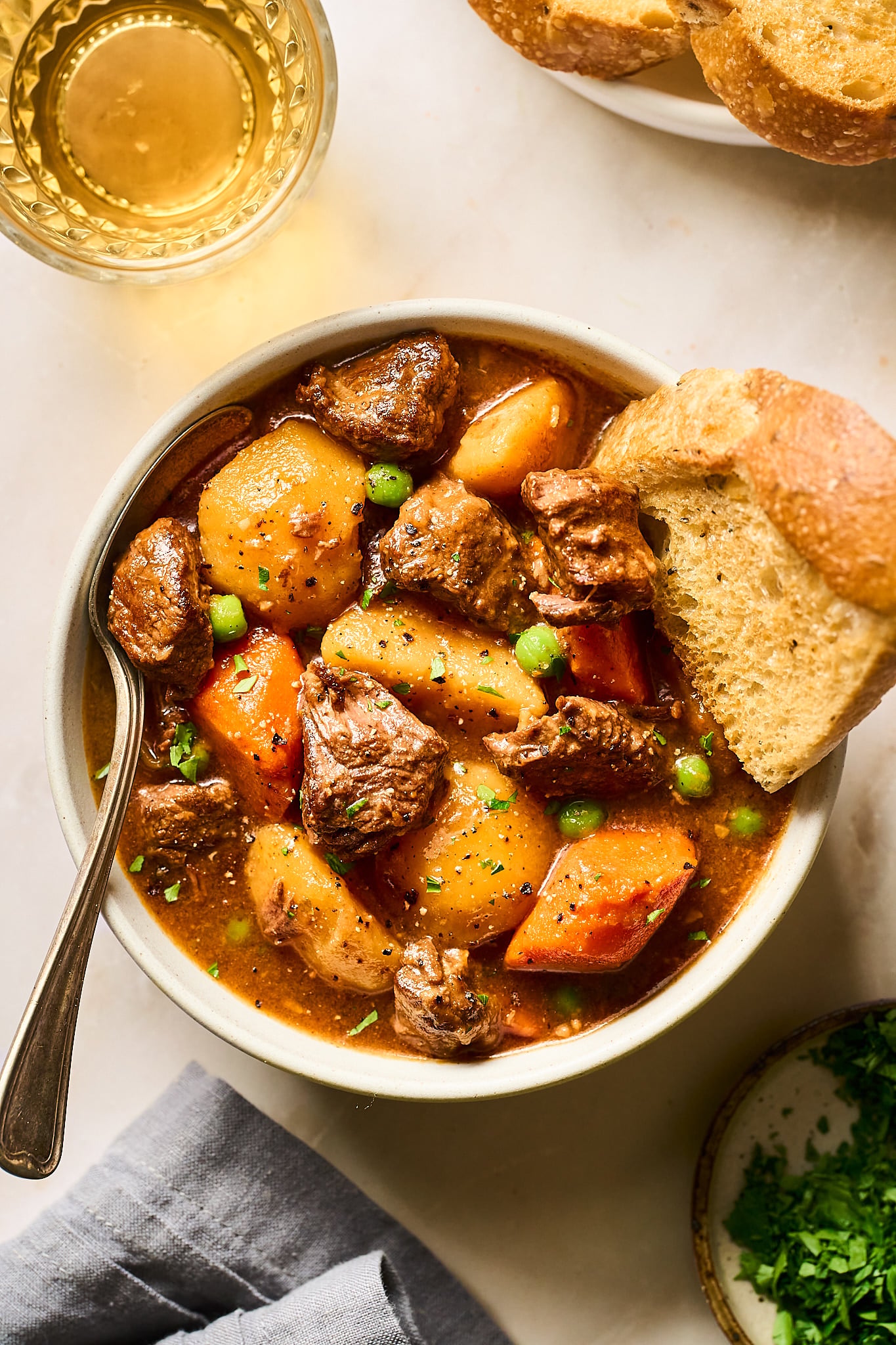 A bowl of beef stew with potatoes, carrots, and peas sitting in a bowl on the countertop. A spoon and a slice of bread also sit in the bowl. Next to the bowl sits a grey cloth napkin, a glass with a slightly yellow, translucent liquid, a plate with two slices of bread, and a plate with chopped parsley.