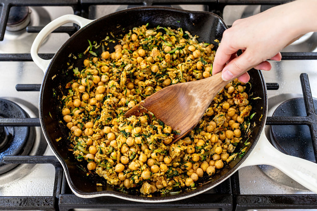 A hand stirs a skillet filled with cooked chickpeas, chopped greens, and spices on a stove, using a wooden spatula. The chickpea mixture appears well-seasoned and sautéed.