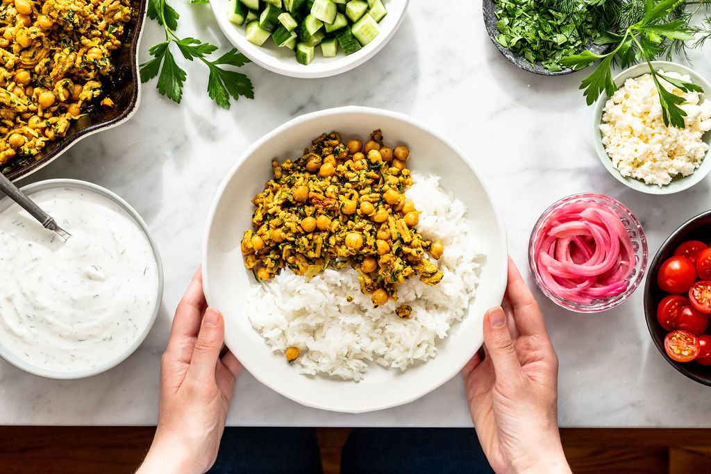 A person holds a bowl of white rice topped with a spiced chickpea and spinach mixture. Surrounding the bowl are bowls of cucumbers, herbs, feta cheese, pickled onions, cherry tomatoes, and a creamy sauce.