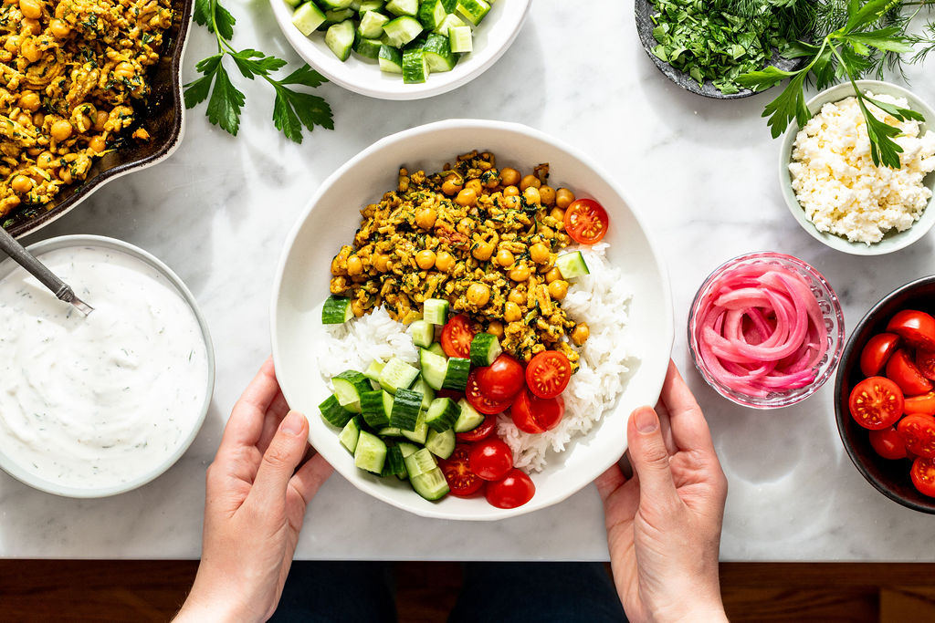 A person holds a bowl of rice topped with spiced chickpeas, chopped cucumbers, and cherry tomatoes. Surrounding bowls contain yogurt sauce, pickled onions, herbs, cheese, more cucumbers, and tomatoes on a white table.