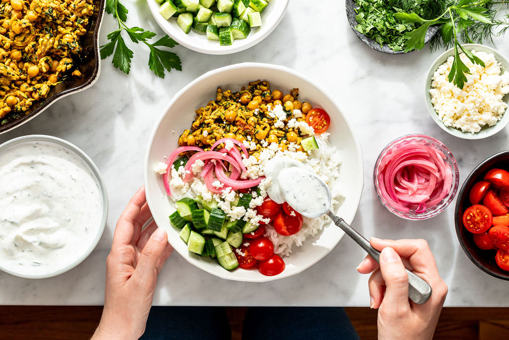 A person holds a bowl of rice topped with cucumbers, cherry tomatoes, pickled onions, and a chickpea mixture, spooning yogurt sauce over it. Surrounding the bowl are fresh herbs, vegetables, and more sauce in bowls.