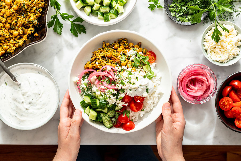 A person holds a bowl filled with rice, cucumbers, tomatoes, pickled onions, herbs, crumbled cheese, and a chickpea mixture, surrounded by bowls of fresh ingredients and sauce on a white countertop.