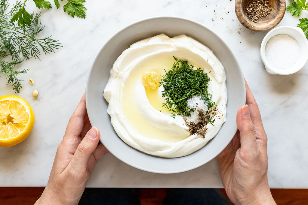 A person holds a bowl of Greek yogurt topped with chopped dill, olive oil, salt, pepper, and a garlic clove, surrounded by fresh herbs, a lemon half, salt, and pepper on a marble surface.