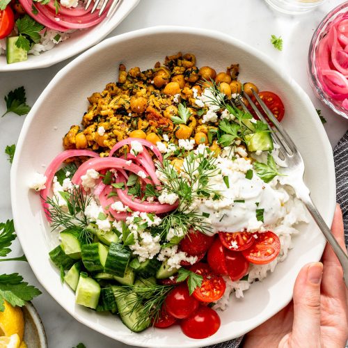 A bowl filled with rice, spiced chickpeas, sliced cucumber, cherry tomatoes, pickled onions, fresh herbs, feta cheese, and a dollop of creamy sauce, with a fork and a hand holding the bowl.