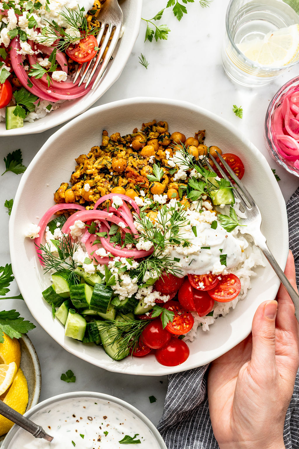 A bowl filled with rice, spiced chickpeas, sliced cucumber, cherry tomatoes, pickled onions, fresh herbs, feta cheese, and a dollop of creamy sauce, with a fork and a hand holding the bowl.