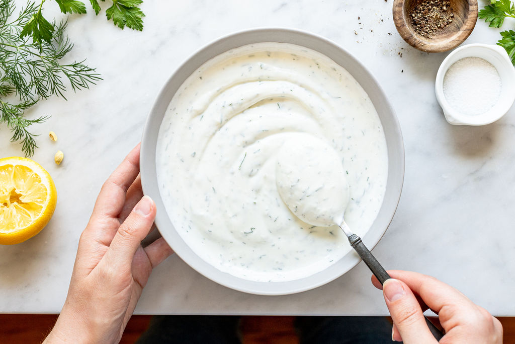 A person holds a bowl of creamy ranch dressing with herbs, stirring it with a spoon. Fresh herbs, a lemon half, salt, and pepper are arranged on a marble countertop nearby.