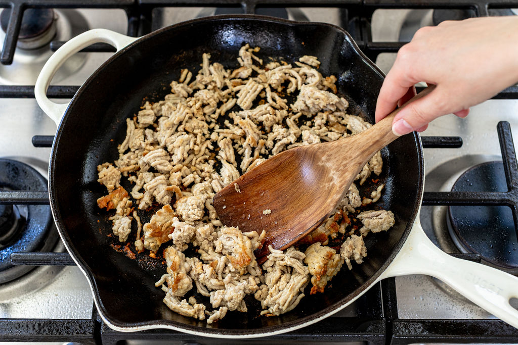 A hand stirs browned ground meat in a black skillet on a gas stove with a wooden spatula.