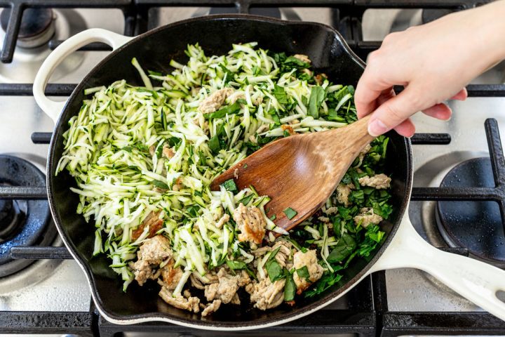 A hand stirs shredded zucchini, chopped greens, and ground meat in a white skillet on a gas stove, using a wooden spoon.