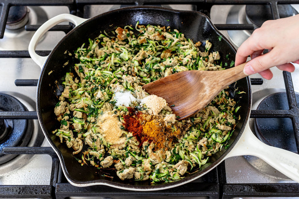 A hand stirs a mixture of shredded zucchini, ground meat, and various spices in a large black skillet on a gas stove.