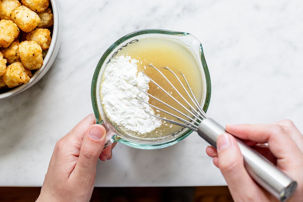 A person whisking cornstarch into a measuring cup of broth on a marble countertop, with a bowl of tater tots nearby.