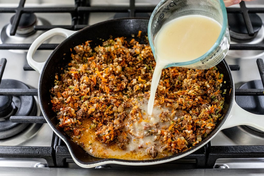 A hand pours broth from a glass measuring cup into a skillet with sautéed ground meat, vegetables, and herbs on a stovetop.