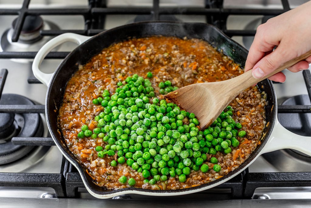 A hand using a wooden spoon to stir frozen green peas into a skillet of simmering ground meat sauce on a stovetop.