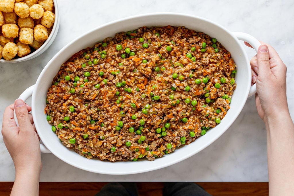 A person holds a white oval dish filled with a mixture of ground meat, peas, carrots, and sauce on a marble surface. A bowl of tater tots is visible nearby.
