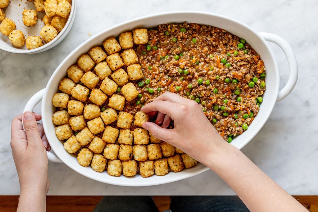 A person arranges tater tots on top of a casserole dish filled with ground meat, peas, and carrots. The dish sits on a marble surface, and a bowl of extra tater tots is nearby.